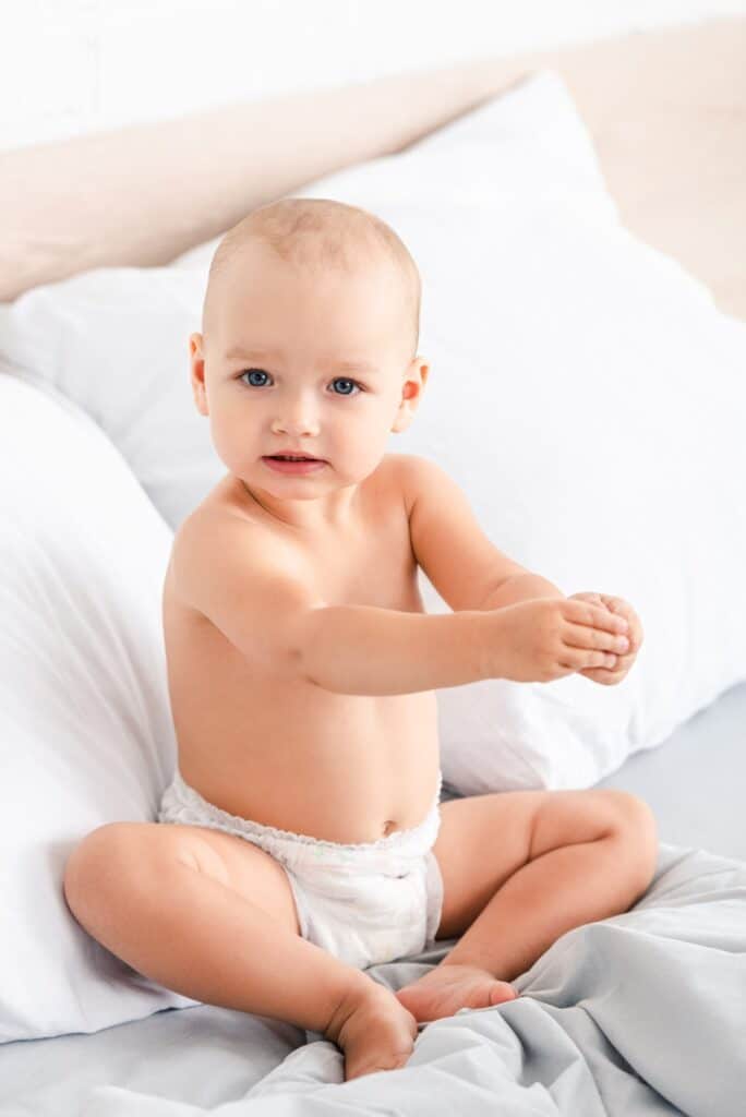 Cute blue-eyed child sitting on bed with white bedclothes and looking at camera